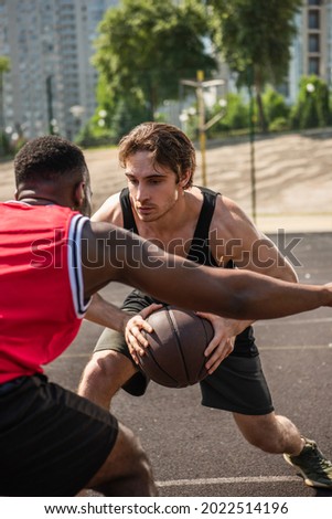 Young sportsman playing basketball with blurred african american friend during match