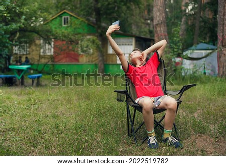 boy is sitting on a folding chair and taking a selfie with a smartphone against the background of summer nature. the photo shows that even in nature and on vacation, children do not part with gadgets