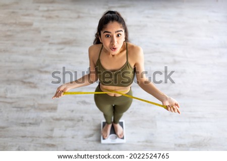Above view of shocked Indian lady standing on scales, checking her waist measurements, surprised with unexpected results of weight loss program, indoors. Slimming diet and workout concept Royalty-Free Stock Photo #2022524765