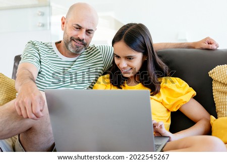 Happy father and teen daughter watching something in her laptop or having a video call sitting in the sofa. Focus on her