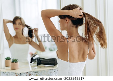 Young confident woman wearing white domestic clothes looking at her reflection while sitting at the mirror and brush long healthy brunette hair. Millennial female doing ponytail at the morning  Royalty-Free Stock Photo #2022568517