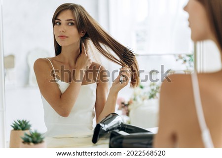 Waist up portrait of the woman reflected in mirror doing daily routine while holding hairbrush tidy her hair. Female looking and tangled hair. Beauty treatment concept 