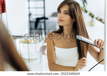 Beautiful young smiling woman using a hair straightener while looking into the mirror in bathroom. Confident girl looking at her reflection with pleasure smile  Royalty-Free Stock Photo #2022568664