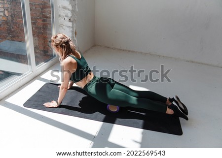 Beautiful and healthy woman wearing green crop top and leggings while lying on massage roller in white studio. Blonde female doing massage on yoga mat with sport equipment.
