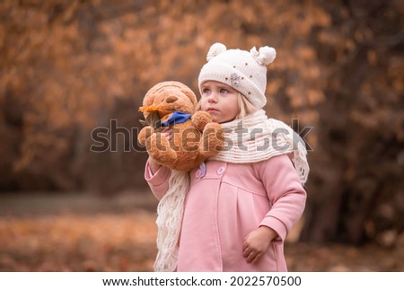 Beautiful little blonde girl, has pretty face, happy emotions, dressed in pink coat, hold soft toy. Child portrait. Lifestyle concept. Autumn time. Fashion kid style.