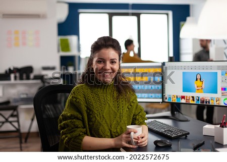 Woman retoucher looking at camera smiling sitting in creative design media agency retouching client photos on PC with two displays. Artist graphic editor working in start up office with digital assets