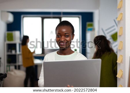 Portrait of african woman looking at camera smiling standing in start up multimedia design agency holding laptop, typing on it. Black video maker editing creative montage using production software