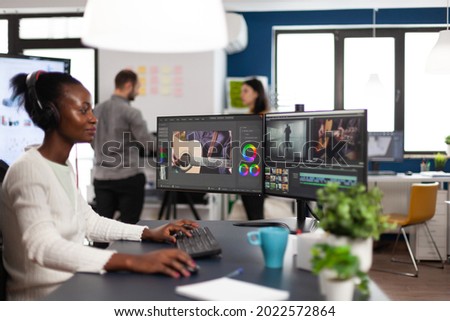 African american video editor wearing headset editing movie using post production software working in start up company office. Black creator processing video footage on computer with dual displays
