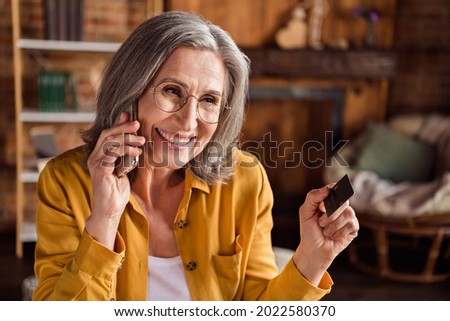 Photo of happy charming woman dressed shirt spectacles talking modern gadget holding credit card indoors workshop workstation