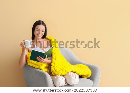 Beautiful young woman with warm plaid drinking tea and reading book while sitting in armchair on color background Royalty-Free Stock Photo #2022587339