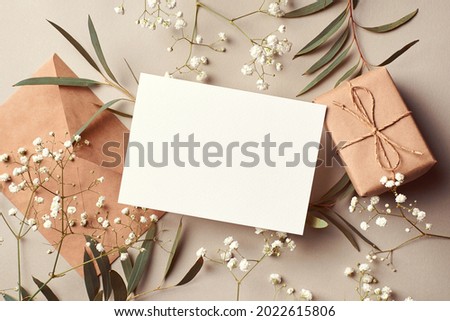 Invitation or greeting card mockup with envelope, gift box and eucalyptus and gypsophila twigs. Card mockup with copy space on beige background.
