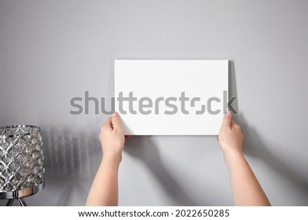 Woman hanging a white canvas mockup on wall. Wall decor. Blank picture in female hands on gray wall background
