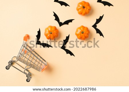 Top view photo of shopping cart model with flying bats silhouettes and small orange pumpkins on isolated beige background with copyspace