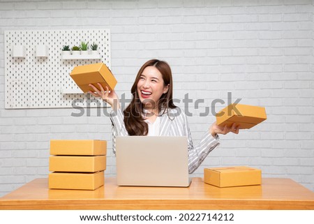 Happy young asian woman startup small business freelance holding parcel box and computer laptop and sitting on chair, Online marketing packing box delivery concept Royalty-Free Stock Photo #2022714212