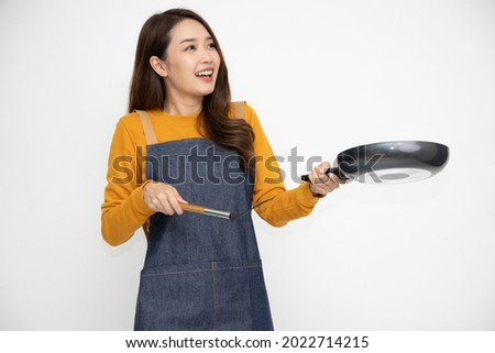 Happy young Asian woman housewife wearing kitchen apron cooking and holding pan and spatula isolated on white background Royalty-Free Stock Photo #2022714215