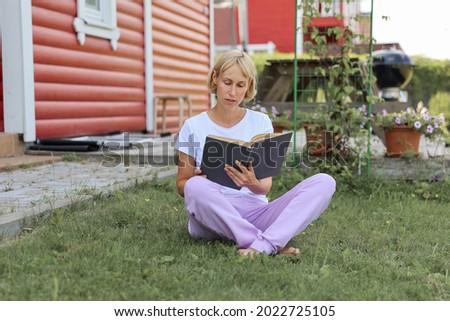 A young woman is sitting on the grass in home yard on a sunny day and reading a real book. Grass and dandilion background, nice vintage look.