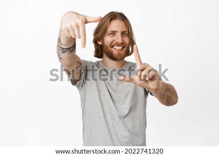 Handsome creative blond guy, smiling and looking through hand frames, picturing, imaging something, creating shooting scene, standing in grey t-shirt over white background