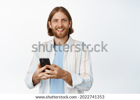 Young man listening music on headphones, smiling, holding mobile phone, using earphones, standing against white background