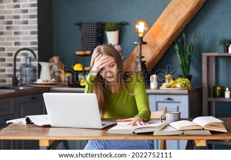 Young pensive woman freelancer sitting at table at home with laptop, making notes, staring thoughtfully into book, thinking about new ideas, female blogger looking for inspiration. Freelance concept
