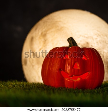 Pumpkin in the form of a head with a creepy smile on the green grass. A lantern lights up inside the pumpkin. There is a large moon in the dark night sky. Halloween. Advertising, trade, banner.