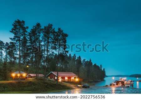 Sweden. Beautiful Red Swedish Wooden Log Cabin House On Rocky Island Coast In Summer Night Evening. Lake Or River Landscape. Beautiful Wooden Pier Near Lake In Summer Evening Night. Lake Or River Royalty-Free Stock Photo #2022756536