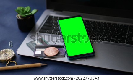 Mobile phone with green screen on a laptop and credit card and bitcoin as background. Concept for freelancing, working from the beach, vacations, holidays, and remote working and home office.
