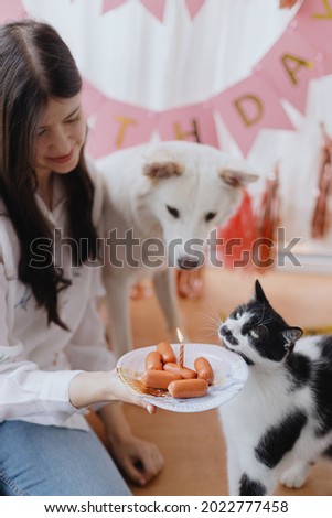 Happy young woman with dog and cat celebrating birthday with sausage cake and candle on background of pink garland in room. Pet birthday party. Adorable dog and cat birthday party at home