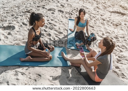 Multiethnic group of female in sports clothing sitting on mat after morning workout, resting and talking. Young athletes sportswomen relaxing after yoga class training together.