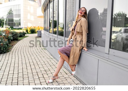 Full length city urban image of fashionable blogger blonde woman wearing beige trench coat, white heeled shoes and trendy accessorizes, posing near business centers, autumn fall time. Royalty-Free Stock Photo #2022820655
