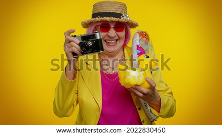 Elderly woman granny tourist photographer taking photos on retro camera and smiling on orange background. Travel, summer holiday vacation. Senior grandmother in sunglasses and hat indoor studio shot