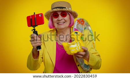 Senior old woman on summer holiday vacation, trip. Mature grandmother traveler blogger with swimming ring and inflatable duck toy taking selfie on mobile phone make video call on orange wall. Tourism