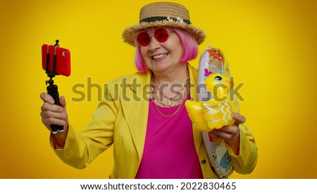 Mature grandmother traveler blogger with swimming ring and inflatable duck toy taking selfie on mobile phone make video call online. Studio shot. Senior woman on summer holiday vacation, trip. Tourism