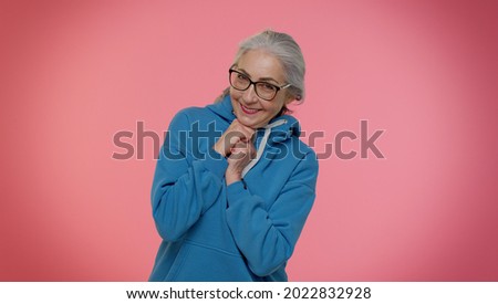 Cheerful lovely senior old gray-haired woman in casual blue hoodie and glasses posing isolated on pink studio wall background. People lifestyle concept. Elderly smiling grandmother looking at camera