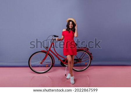 Full-length portrait of graceful girl in summer red dress standing in studio on purple background. Carefree brunette woman in straw hat posing with her bicycle.