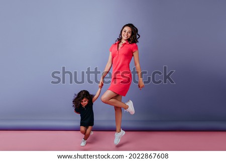 Full-length portrait of stunning young mom holding hands with her kid. Spectacular brunette woman dancing with daughter on purple background.