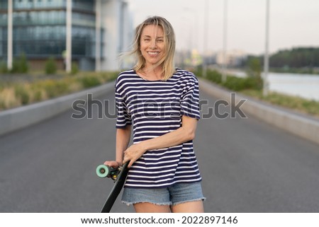 Overjoyed woman of middle age happy after skateboarding. Portrait of cheerful female of 40s hold longboard. Active skateboarding lady enjoy skating in summer evening on empty road. Lifestyle concept