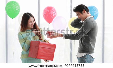 Pretty young Asian woman happily smile and glad getting lovely red gift box from beloved boyfriend at romantic indoor party decorated by fancy balloon to celebrate ecstatic love in winter
