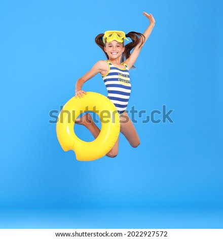 Little funny happy girl in swimsuit and goggles on her head jumping up in air with inflatable ring around waist, isolated over blue studio background. Summertime, vacation an school holidays concept Royalty-Free Stock Photo #2022927572