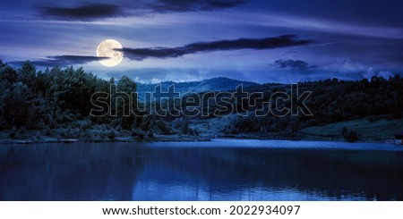 lake among mountain landscape in spring at night. beautiful countryside scenery with forest on the shore in full moon light. clouds on the sky
