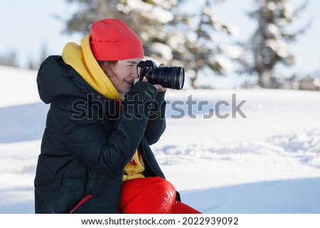 Woman photographer in ski equipment makes pictures sitting in the snow. Holds a camera with a huge lens. Winter clothing: pants and warm jacket, knitted hat, ski goggles. Selective focus.