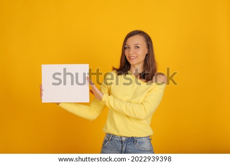 a young beautiful blonde girl in a yellow jacket and jeans holds a white sheet of paper in her hands