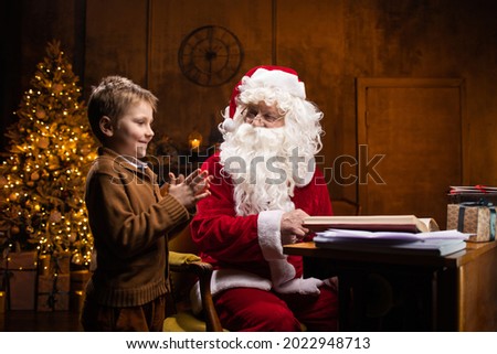 Santa Claus and little boy. Cheerful Santa is working while sitting at the table. Fireplace and Christmas Tree in the background. Christmas concept.