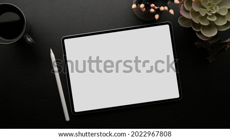 Portable tablet white screen mockup with stylus pen on dark working space with house plant, pencils, coffee, black background, overhead