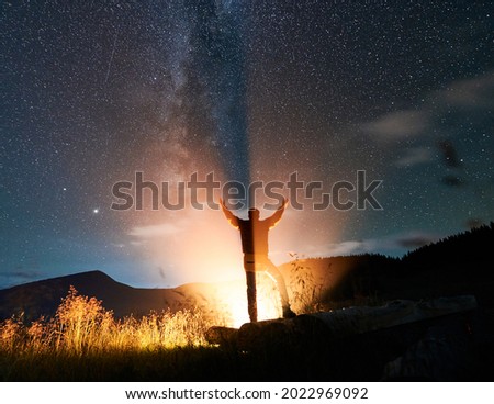 Back view of man hiker looking at night starry sky and raising hands while bonfire illuminating male silhouette. Traveler reaching out his hands to night sky with stars. Concept of travelling.