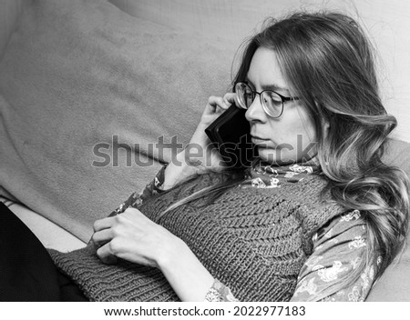Upset young woman with smartphone in her hands talking on phone while sitting on couch at home. Black and white photo.