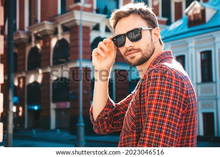 Handsome smiling stylish hipster lambersexual model.Modern man dressed in red checkered shirt. Fashion male posing near skyscraper on the street background in sunglasses. Outdoors at sunset 