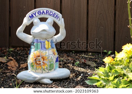 An adorable, stone lawn ornament in the shape of a frog. There are tile letters on the top that read "MY GARDEN". The frogs belly has tiles on it as well that make out the shape of a sunflower.
