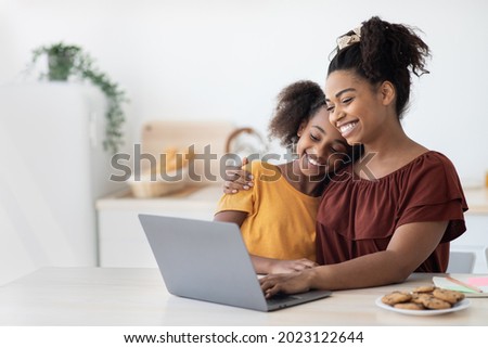 Cute black mother and kid using laptop together, sitting at kitchen table with homemade pastry, hugging and looking at computer screen, spending time together, watching cartoon, copy space