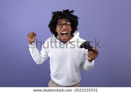 Triumphant black teen guy with joystick shouting in excitement, playing video game, making YES gesture on violet background. Overjoyed teenage gamer with controller winning online gaming championship
