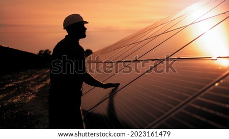 Assistance technical worker in uniform is checking an operation and efficiency performance of photovoltaic solar panels. Unidentified solar power engineer touches solar panels with his hand at sunset Royalty-Free Stock Photo #2023161479